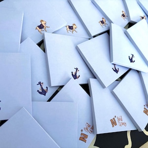 May include: A collection of white square notepads with various nautical-themed illustrations. Some pads feature a running horse, while others display a navy blue anchor. The phrase "N Beat Army" is printed in orange on some pads.