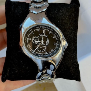 Könnte beinhalten: Eine silberfarbene Denvosi Quarzuhr mit schwarzem Zifferblatt, ausgestattet mit mehreren Hilfszifferblättern, einer Datumsanzeige und Monatsindikatoren. Die Uhr hat ein poliertes, unregelmäßig geformtes Metallgehäuse und ein passendes Gliederarmband.