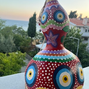 May include: A hand-painted gourd with a bulbous base and a narrower neck, featuring a vibrant red background decorated with multiple blue, white, and yellow eye motifs, white polka dots, a purple flower, and colourful wavy horizontal stripes.