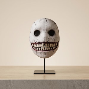 Könnte beinhalten: Eine weiße, ovale Maske mit tiefen, dunklen Augenhöhlen und einem breiten, zahnigen Grinsen mit sichtbaren roten Details um das Zahnfleisch. Die Oberfläche weist feine Risse und verwitterte Texturen auf, montiert auf einem dünnen schwarzen Metallständer.