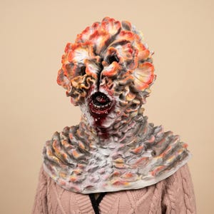 Op de afbeelding: Een masker voor het hele hoofd met een gestructureerd, op schimmels geïnspireerd ontwerp met gelaagde, gegolfde uitsteeksels in tinten oranje, rood en grijs. Het masker heeft een centrale verticale spleet die een donkere, open mond onthult.