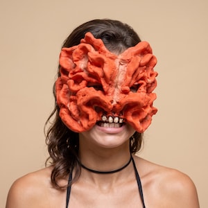 Op de afbeelding: Een fel oranje, getextureerd masker met een complexe, gevouwen, organische oppervlaktestructuur. Het masker heeft een rij kleine, gebroken witte, rechthoekige tanden in het midden, die het bovenste deel van het gezicht bedekken.