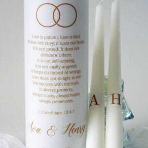 May include: A white unity candle and two taper candles for a wedding. The unity candle has gold rings and text from 1 Corinthians 13:4-7. The taper candles are initialed with "A" and "H". The wedding date is April 12, 2028.