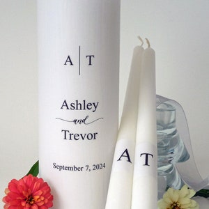 May include: A white pillar candle and two taper candles with the initials "A T" and the names "Ashley and Trevor" and the date "September 7, 2024" printed on them. A red and yellow flower are in the foreground.