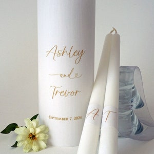 May include: A white pillar candle and two taper candles with gold lettering. The pillar candle is inscribed with "Ashley and Trevor" and the date "September 7, 2024." The taper candles have the initials "AT."