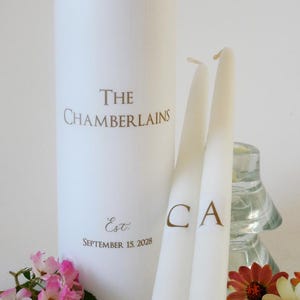 May include: A large white pillar candle printed with The Chamberlains and Est. September 15, 2028 in gold lettering, accompanied by two slim white taper candles featuring the gold initials C and A, decorated with small pink and orange flowers.