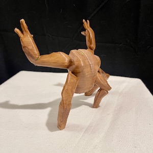 May include: A brown wooden sculpture of a figure in a yoga pose. The figure has visible wood grain and is posed with arms raised and legs bent. The sculpture is on a white surface against a black backdrop.