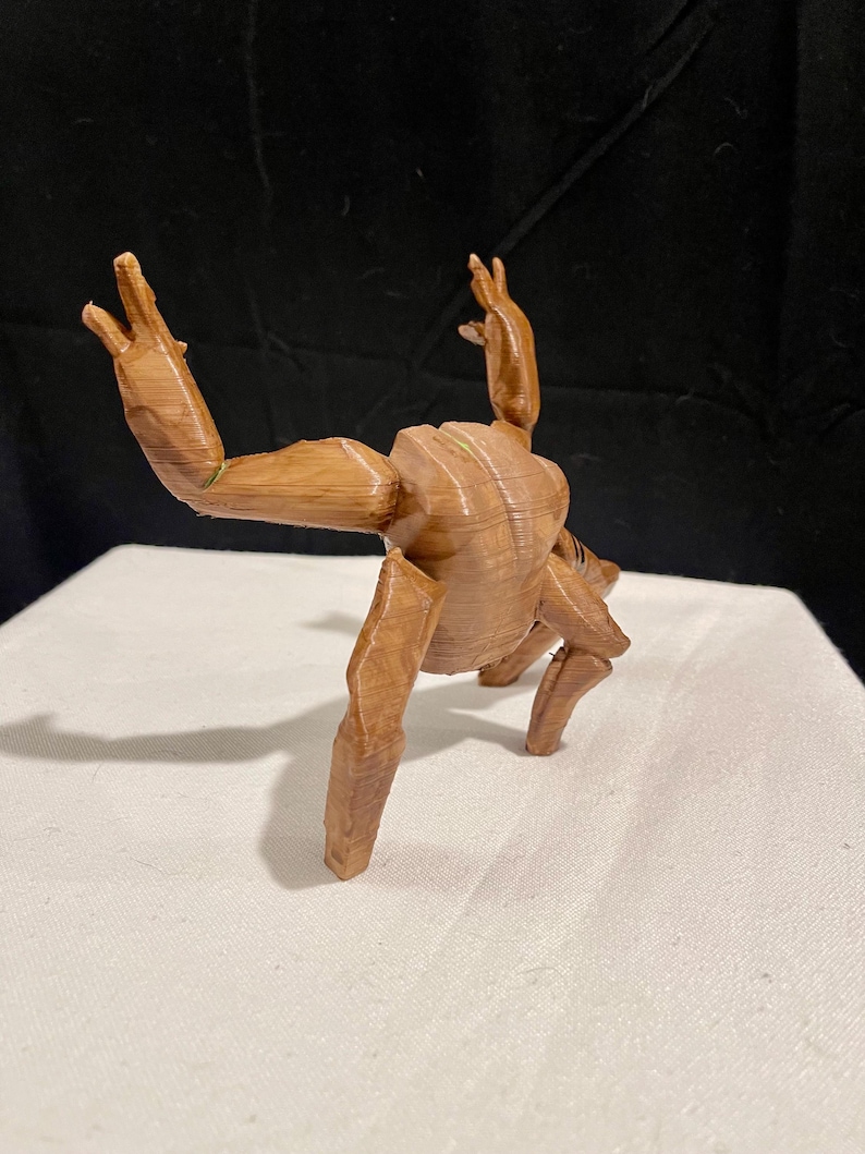 May include: A brown wooden sculpture of a figure in a yoga pose. The figure has visible wood grain and is posed with arms raised and legs bent. The sculpture is on a white surface against a black backdrop.