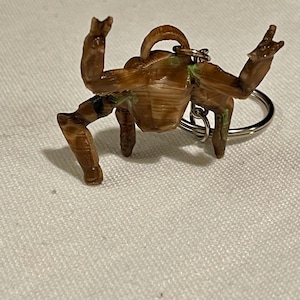 May include: A brown crab-shaped keyring with a silver ring. The crab has its arms raised and legs extended. Green paint accents the joints. The keyring is made of a textured material.