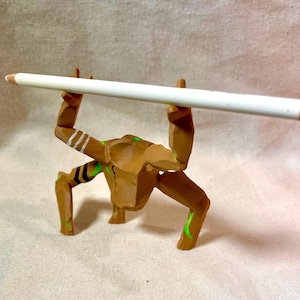 May include: A brown wooden figurine supporting a white pencil with a red end. The figurine has green and white details. The pencil is held horizontally, creating a unique display.