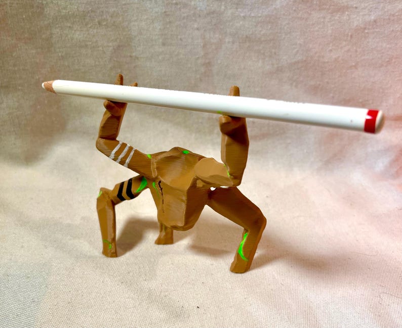 May include: A brown wooden figurine supporting a white pencil with a red end. The figurine has green and white details. The pencil is held horizontally, creating a unique display.