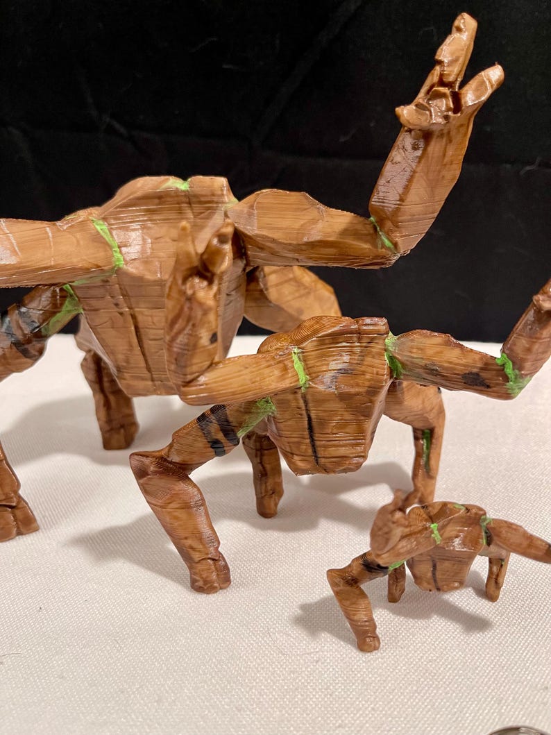 May include: Three wooden sculptures of a creature with outstretched arms and legs. The sculptures are brown with green accents and black stripes. The largest sculpture is in the centre, with two smaller ones on either side.
