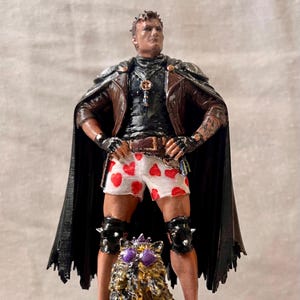 May include: A hand-painted figurine of a person wearing a brown leather jacket, a black shirt, and white shorts with a red heart pattern. The figure wears a black cape, fingerless gloves, and black knee pads with spikes, standing behind a small creature.