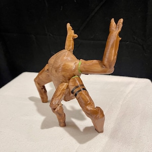 May include: A wooden figurine of a creature with outstretched arms and legs. The figure is brown with black stripes on its legs and a green band around its torso. The figurine is posed on a white surface against a black backdrop.