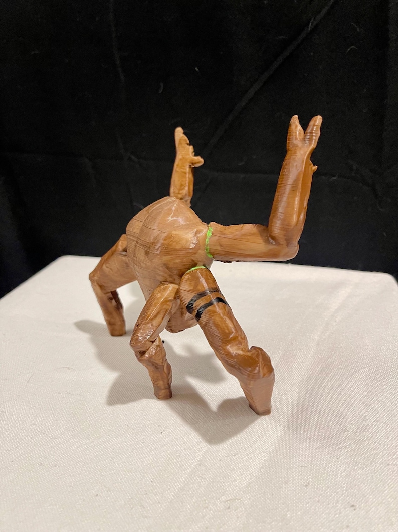 May include: A wooden figurine of a creature with outstretched arms and legs. The figure is brown with black stripes on its legs and a green band around its torso. The figurine is posed on a white surface against a black backdrop.