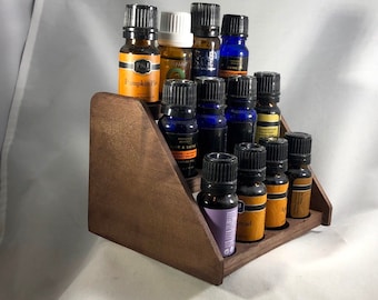 Puede incluir: Un expositor de aceites esenciales de madera en niveles. El soporte marrón oscuro contiene varias botellas de vidrio pequeñas con tapas negras. Las botellas contienen varios aceites esenciales, con etiquetas visibles. El texto "Wood finish" está en la parte inferior de la imagen.
