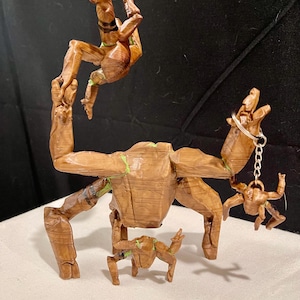 May include: A wooden sculpture of a tree-like figure with outstretched arms, holding a small keychain figure. Another figure is suspended above. The sculpture is brown with green accents.