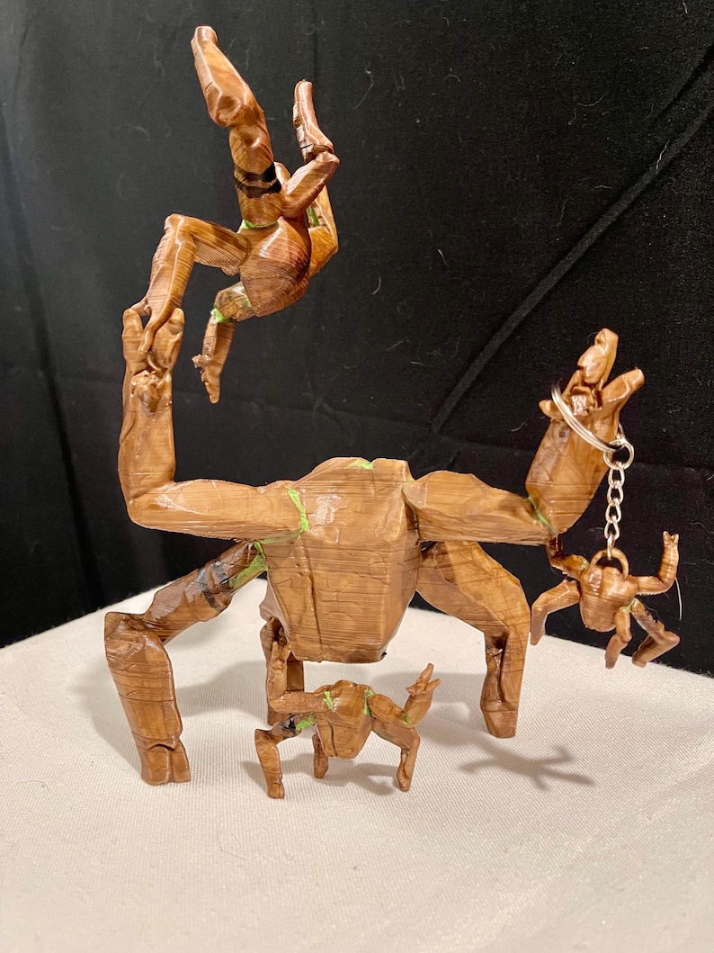 May include: A wooden sculpture of a tree-like figure with outstretched arms, holding a small keychain figure. Another figure is suspended above. The sculpture is brown with green accents.