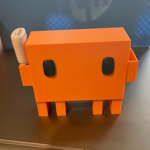 May include: An orange, geometric, block-shaped toy with black oval eyes and four square legs. A white cylindrical piece is attached to the side. The toy is designed with a minimalist aesthetic.