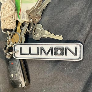 May include: A white and black keychain with the word "LUMON" printed on it. The keychain is attached to a set of keys.