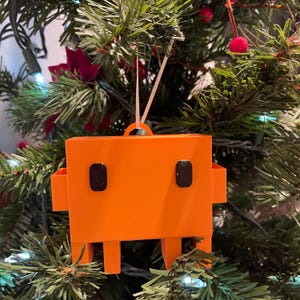 May include: A bright orange, rectangular, blocky ornament with two small black square eyes and two side protrusions, hanging from a white string. The object has a flat top with a small loop for hanging and four short legs at the bottom base.