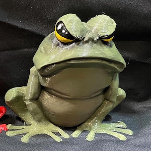 May include: A 3D printed green frog figurine with yellow eyes. The frog has a grumpy expression and is sitting on a black surface.