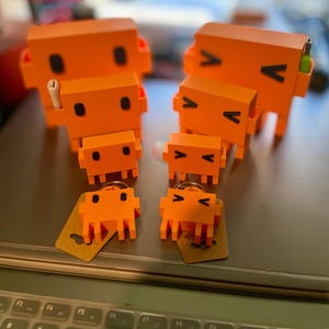 May include: A collection of orange, cuboid robot-style figures in different sizes. Some have black V-shaped markings and small black dots. Two are keyrings with tags. One larger figure has a white cylindrical object inserted.