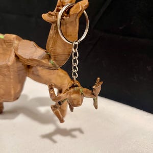 May include: A brown 3D-printed keychain with a small figure with outstretched arms, attached to a silver keyring and chain. The keychain is a miniature version of a larger wooden sculpture, with visible details and textures.