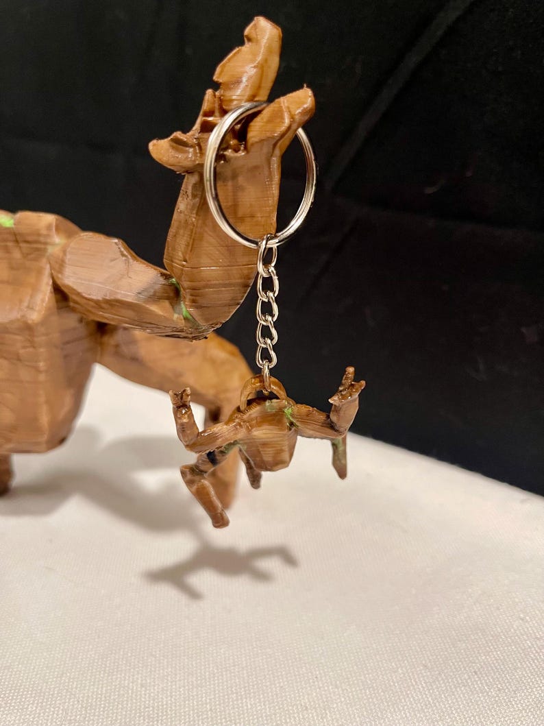 May include: A brown 3D-printed keychain with a small figure with outstretched arms, attached to a silver keyring and chain. The keychain is a miniature version of a larger wooden sculpture, with visible details and textures.