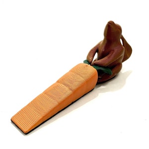 May include: A brown rabbit figurine holding a carrot-shaped doorstop. The carrot is orange and has a green stem.