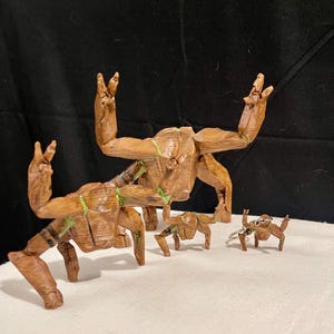 May include: A set of wooden Groot figures in different sizes. The figures are brown with green detailing and are posed with their arms raised in a peace sign. They are on a white surface with a black backdrop.