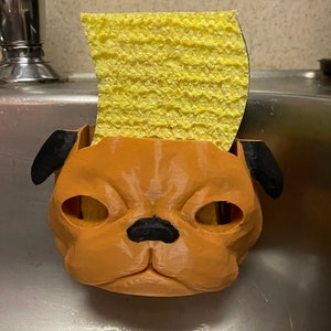 May include: A 3D printed brown pug-shaped sponge holder with black ears and a black nose. A yellow sponge is placed inside the holder.