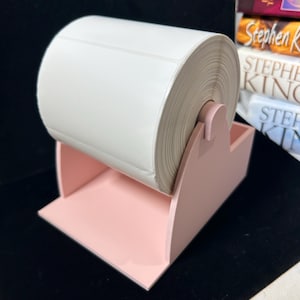 Puede incluir: Un soporte de plástico rosa claro para rollos de etiquetas de escritorio con una estructura de soporte curva que sostiene un gran rollo de etiquetas adhesivas rectangulares blancas en blanco, con base abierta y eje central.