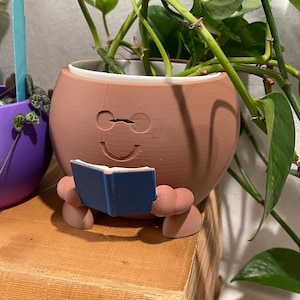 May include: A terracotta-colored, round planter with a smiling face design, holding a blue book. The planter is filled with green leafy plants. A purple planter is visible to the left. The planter is sitting on a brown surface.