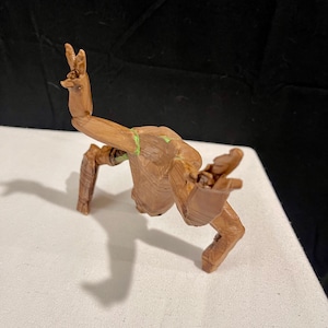 May include: A brown, articulated figure of a character with a tree-like appearance, posed with arms raised in a peace sign. The figure is on a white surface, with a black background.
