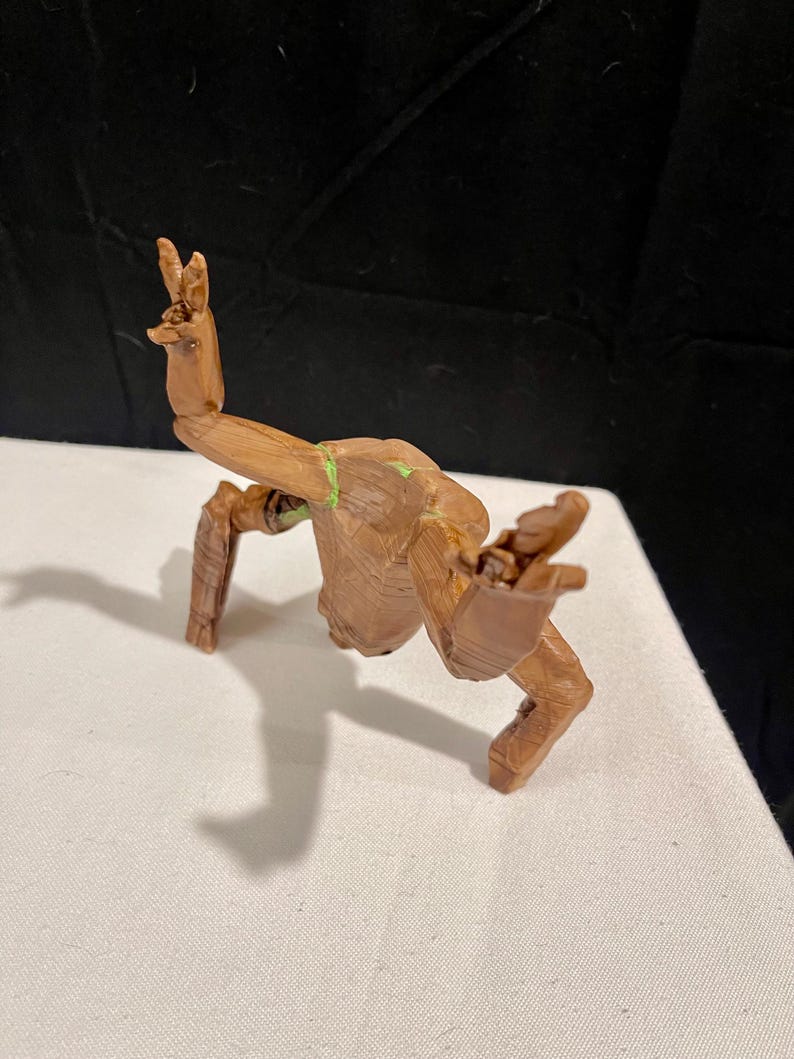 May include: A brown, articulated figure of a character with a tree-like appearance, posed with arms raised in a peace sign. The figure is on a white surface, with a black background.