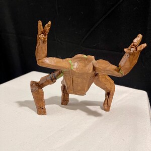 May include: A wooden sculpture of a character with outstretched arms and legs. The figure is brown with green accents and appears to be carved from wood. The sculpture is posed on a white surface against a dark background.