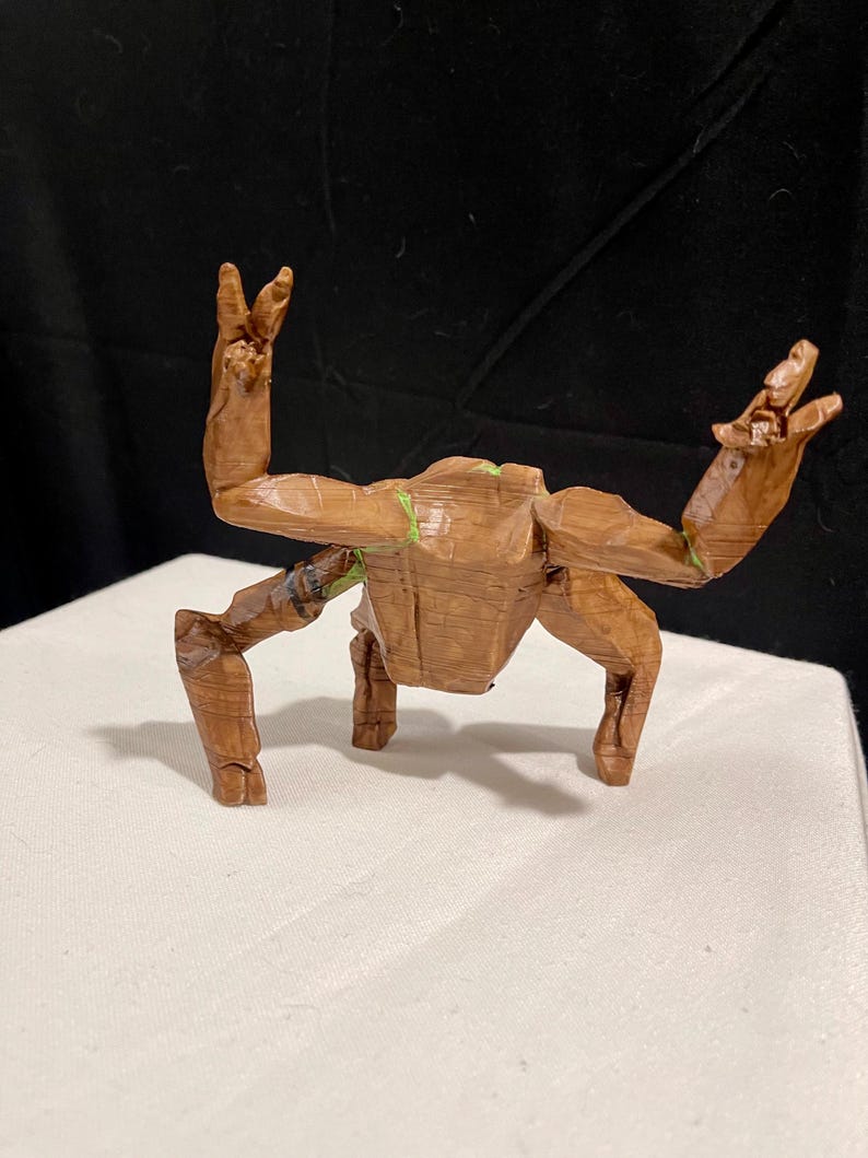 May include: A wooden sculpture of a character with outstretched arms and legs. The figure is brown with green accents and appears to be carved from wood. The sculpture is posed on a white surface against a dark background.