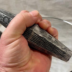 May include: A dark grey, multi-faceted, tapered object with a textured, wood-like grain finish. The top section features a circular emblem with a cross-like pattern. The object has a geometric, elongated shape with distinct edges and a weathered appearance.