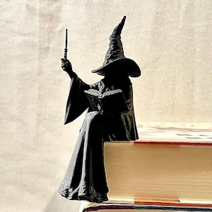 May include: A black figurine of a robed wizard wearing a pointed hat, sitting on the edge of a book. The wizard holds a thin wand in one hand and an open book in the other, with a textured surface finish visible on the dark material of the figure.