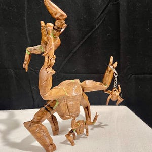 May include: A wooden sculpture of a character with a tree-like appearance, featuring multiple articulated limbs and a small figure hanging from a chain. The sculpture is primarily brown with green and black accents, and is posed on a white surface.