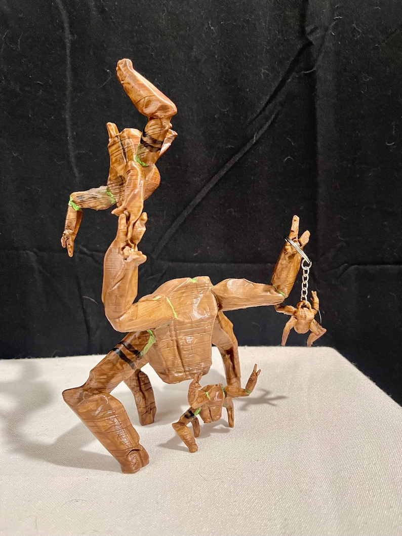 May include: A wooden sculpture of a character with a tree-like appearance, featuring multiple articulated limbs and a small figure hanging from a chain. The sculpture is primarily brown with green and black accents, and is posed on a white surface.