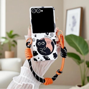 May include: A white foldable smartphone case featuring a black cat illustration, the text "BLACK CAT" and "Good night", and various small motifs like bats and stars. The case includes an attached long, adjustable orange and black rope lanyard.