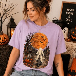Pode incluir: Uma t-shirt roxo-claro com uma estampa gráfica de um fantasma andando de bicicleta numa rua à noite. A cena inclui uma grande lua cheia laranja, árvores sem folhas, casas, uma cerca branca e abóboras sob um céu com morcegos a voar.