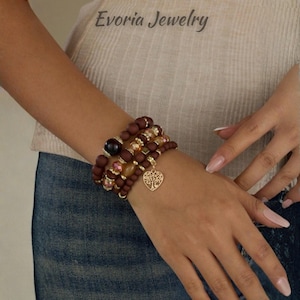 Pode incluir: Um conjunto de pulseiras de várias voltas com contas castanhas mate, contas facetadas cor de âmbar e pequenos espaçadores metálicos dourados. Um pendente dourado em forma de coração com um design recortado de árvore da vida pende do conjunto.
