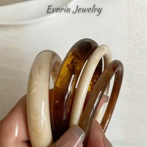 May include: A set of four circular bangles featuring a smooth, polished finish. The bracelets alternate between a solid cream colour and a translucent, deep amber brown tone. The text Evoria Jewelry is visible at the top of the image.