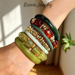 Könnte beinhalten: Ein Stapel aus sechs Armbändern mit einer Mischung aus glatten, olivgrünen, durchscheinend hellgrünen, blassminzfarbenen und dunkelwaldgrünen Harzreifen sowie goldfarbenen Metallperlen und dunkel bernsteinfarbenen Harzsegmenten von Evoria Jewelry.