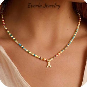 May include: A beaded necklace featuring small, round, metallic gold-tone beads interspersed with tiny, colourful, multi-hued beads. A gold-tone letter A charm hangs from the centre of the necklace. The text Evoria Jewelry is visible at the top.