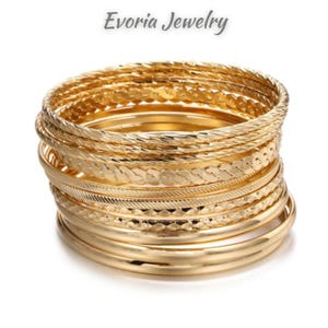 Könnte beinhalten: Ein Stapel goldfarbener Metallarmreifen mit verschiedenen Texturen, darunter gedrehte, geflochtene und glatt polierte Oberflächen. Der Text Evoria Jewelry wird über der Sammlung kreisförmiger Armbänder angezeigt.