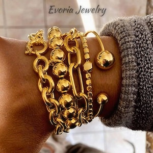 May include: A collection of gold-toned bracelets featuring a chunky oval link chain, a strand of polished spherical beads, a delicate beaded chain with a hexagonal accent, and an open cuff bracelet with two large spherical ends. Text reads Evoria Jewelry.